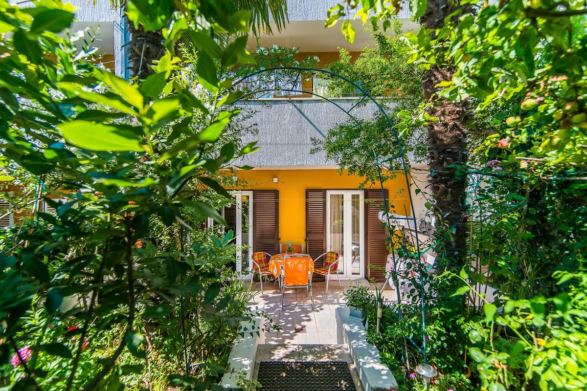 A welcoming terrace is seen through a lush garden archway, featuring seating furniture in bright orange. The entrance to the apartment is flanked by green foliage, with dual glass doors that invite light while being sheltered by wooden shutters.