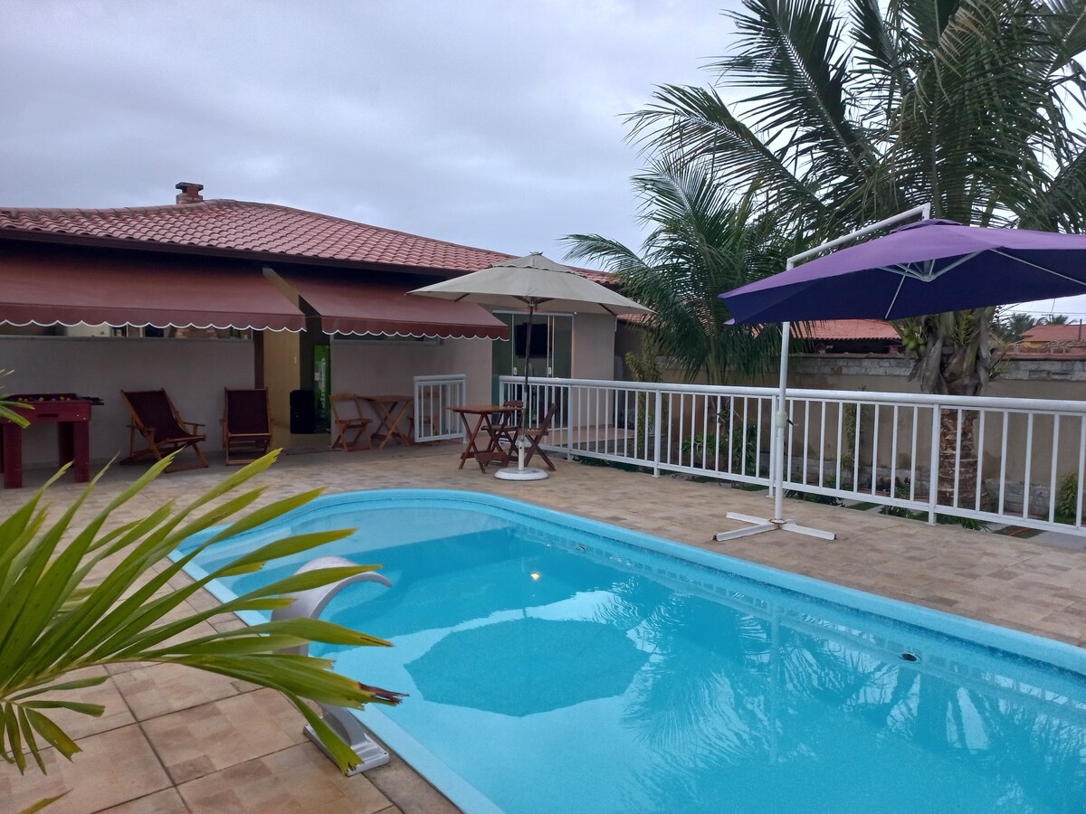 A refreshing swimming pool is centered within a spacious outdoor area, surrounded by lounging chairs and umbrellas. Tropical palm trees provide shade, while covered seating areas invite relaxation. The overall setting conveys a tranquil environment for family activities and leisure.