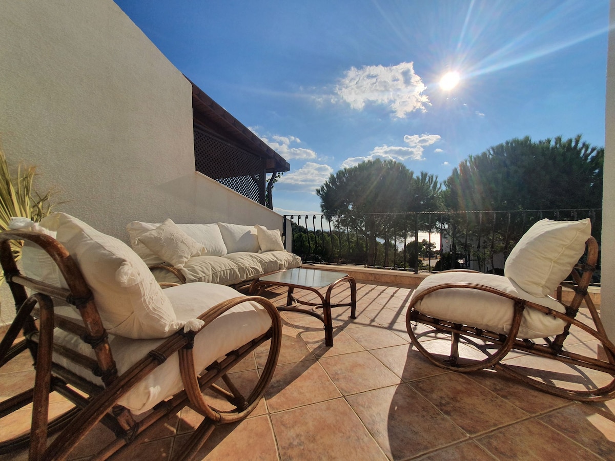 An inviting terrace area features two rocking chairs and a glass-top table, all surrounded by tile flooring. The sun shines brightly overhead, illuminating the lush greenery in the background. The view opens up towards the sea, enhancing the serene ambiance.