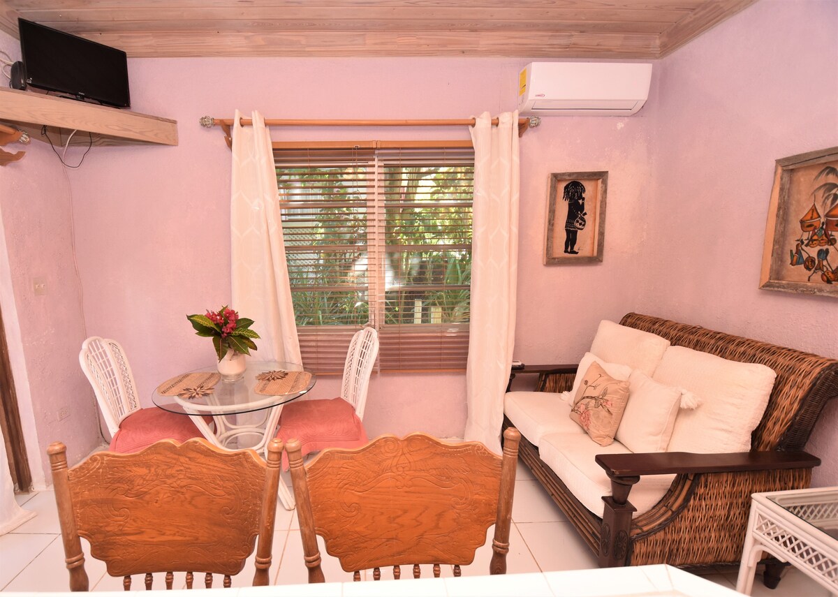 A cozy seating area is furnished with a wicker sofa and a glass dining table surrounded by four wooden chairs. Natural light filters through a window adorned with sheer curtains, highlighting pastel purple walls and a decorative wall piece.