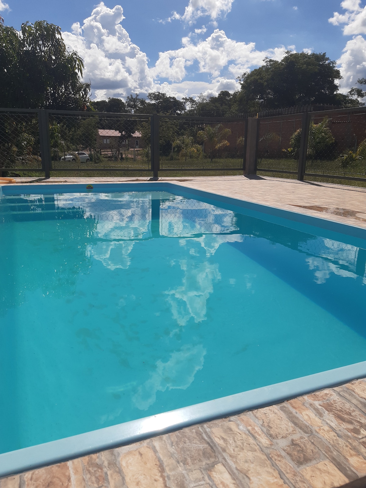 A clear swimming pool reflects the clouds above, surrounded by a secure fence. The pool area is complemented by a tiled deck, offering a serene view of the lush greenery and distant structures in the background.