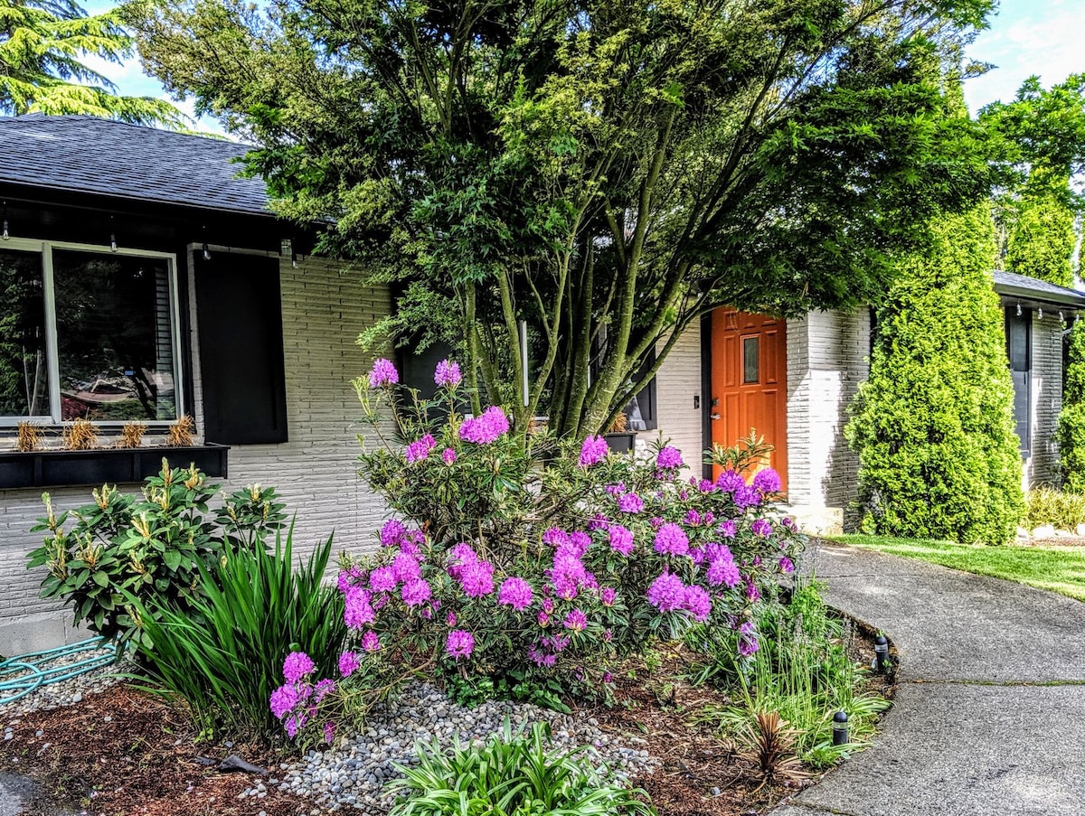 A well-maintained front yard features vibrant purple flowers blooming among lush greenery. A curved pathway leads to a welcoming entrance, which is highlighted by a bright orange door, framed by mature trees and neatly arranged shrubs.