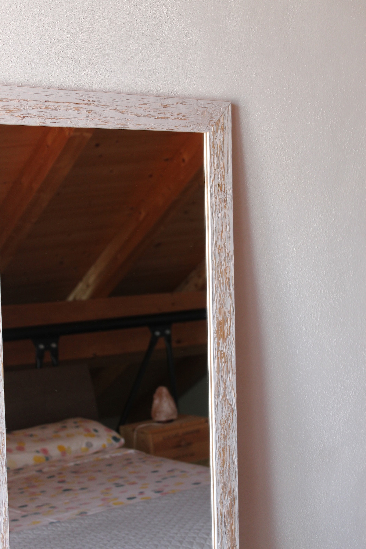 A tall mirror with a weathered frame is positioned against a light-colored wall, reflecting part of a cozy sleeping area. The bed features a neutral bedspread with a subtle pattern, and wooden beams of the ceiling add warmth to the space.
