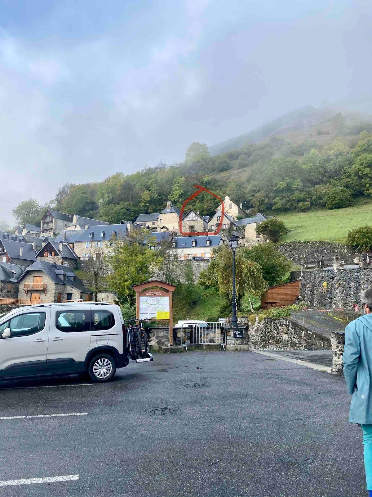 A charming hamlet is nestled in a mountainous area, with several stone buildings visible on a hillside. Green vegetation covers parts of the surrounding area, and a vehicle is parked in the foreground. Low-lying clouds partially obscure the hilltop.