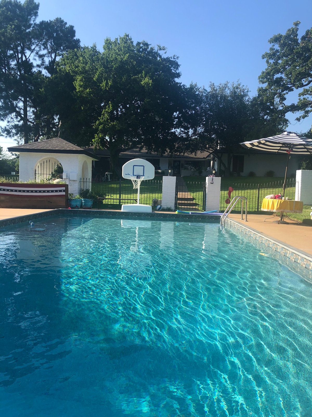 A clear blue pool is surrounded by a spacious deck, featuring a basketball hoop installed at one end. Lush green trees and a shaded gazebo are visible in the background, adding to the outdoor ambiance. Bright sunlight reflects off the water’s surface.