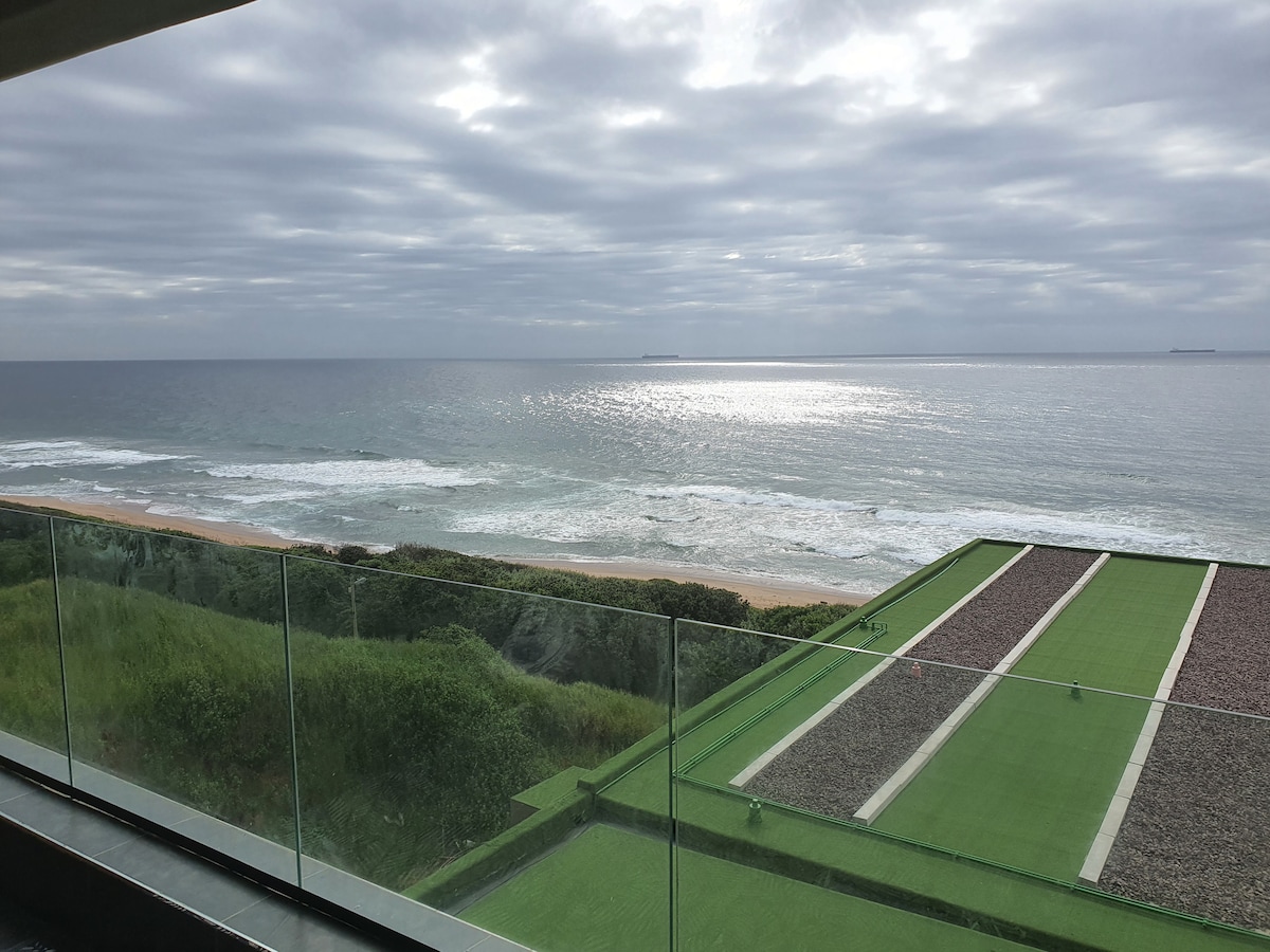 A panoramic view showcases the ocean's tranquil waves crashing against the shore. Verdant foliage lines the beach, while a spacious terrace with glass panels allows for unobstructed sightlines of the horizon and the shimmering water under a cloudy sky.