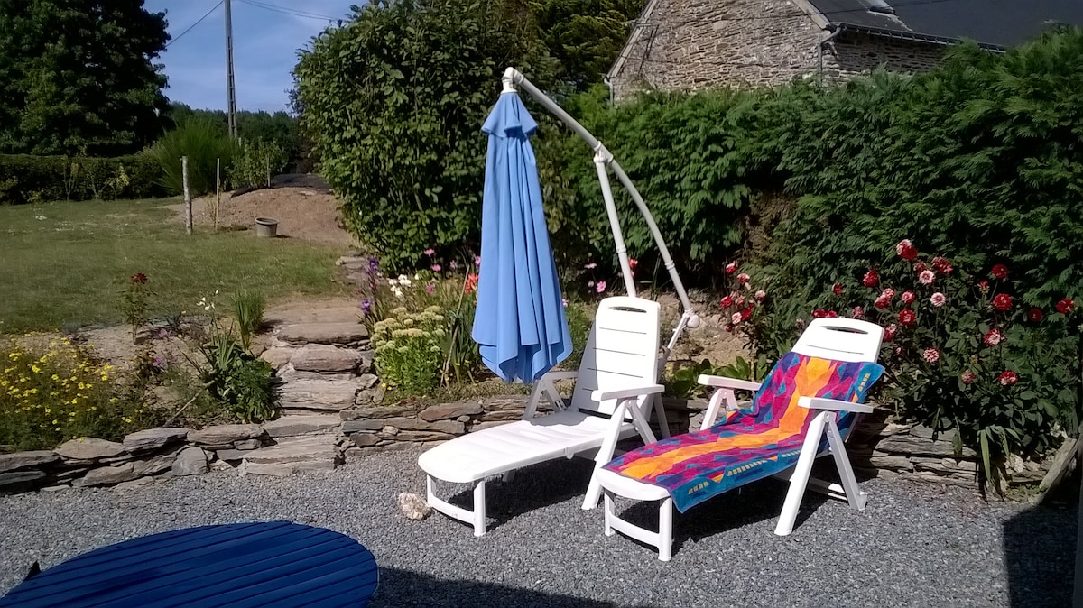 A private gravelled area features two lounge chairs, one draped with a colorful towel. A blue umbrella is positioned beside the chairs, providing shade. Surrounding greenery includes flowering plants and shrubbery, creating a tranquil outdoor space.