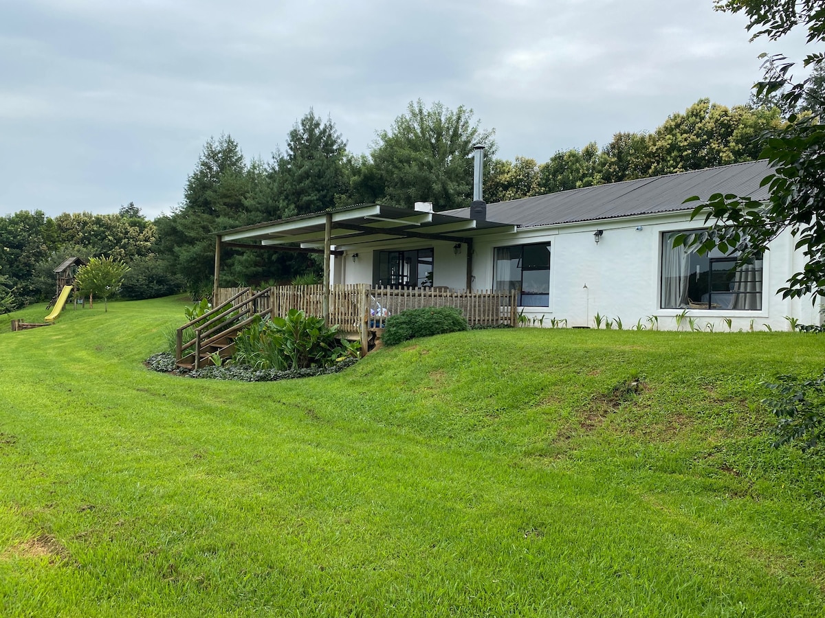 The cottage is nestled amidst lush greenery, showcasing a well-maintained lawn. A spacious covered patio extends from the cottage, with views of the garden and play area, highlighted by a slide in the background. Large windows bring natural light into the living spaces.