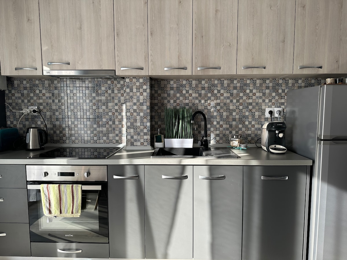 The kitchen is equipped with modern appliances including an oven, a stovetop, and a refrigerator. Square tiles create a mosaic backsplash, while a sink is situated beneath a window. Greenery is displayed on the countertop, adding a touch of freshness to the space.