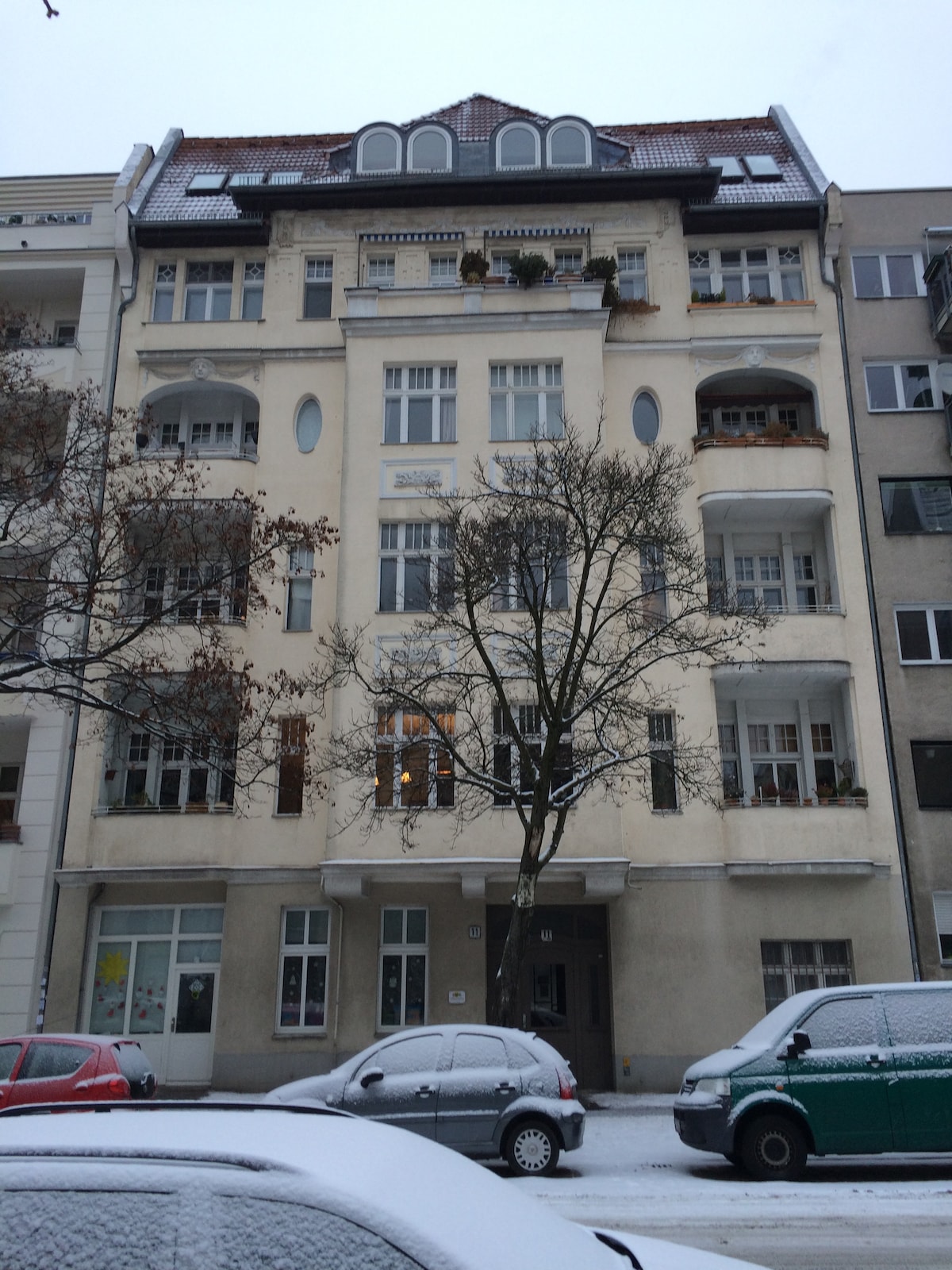 A historic building built in 1906 is showcased, featuring a decorative facade with multiple windows and balconies. The structure presents a symmetrical design, highlighted by arched windows and ornate detailing. A light dusting of snow covers the ground and nearby vehicles, adding a tranquil ambiance.