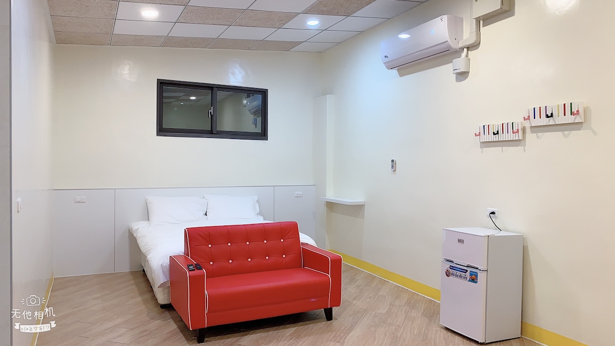 A bright, minimalist room is featured, with a spacious bed covered in white linens. A red couch is positioned near a small white refrigerator. A window allows natural light into the room, while air conditioning is provided on the wall.