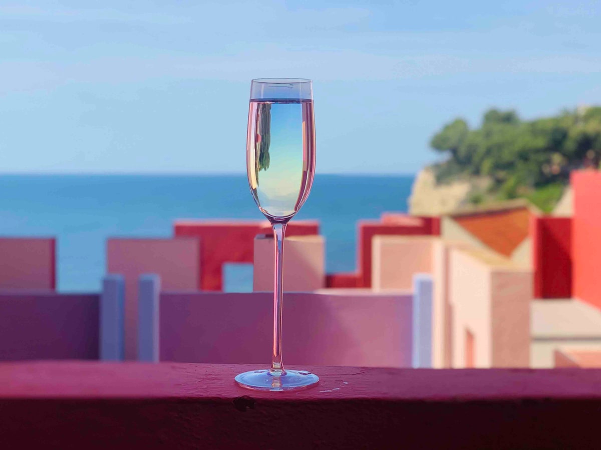A glass of sparkling drink is placed on a ledge, with the sea visible in the background. Colorful seaside structures in various pastel shades complement the view, creating a vibrant and serene coastal atmosphere.