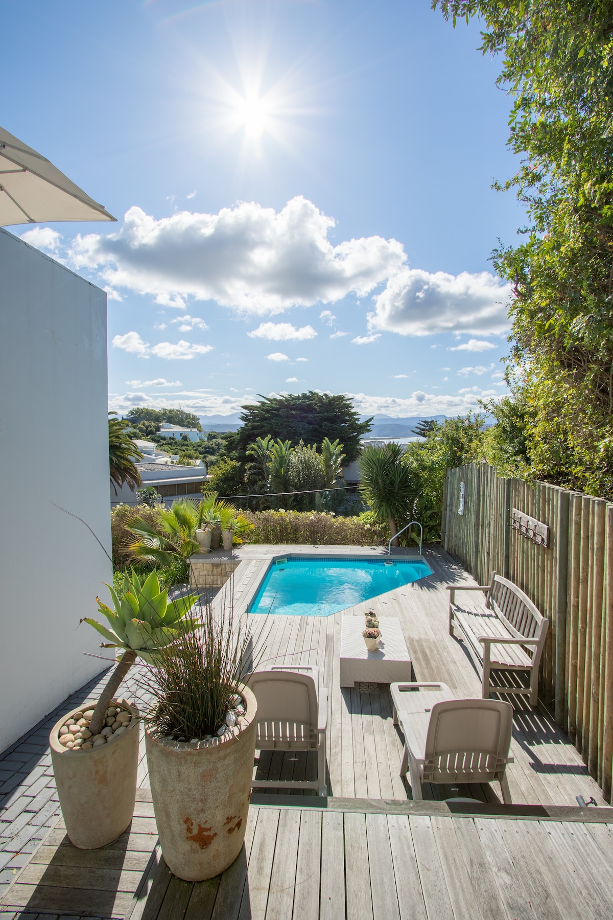 A serene outdoor space features a private plunge pool surrounded by wooden decking. Comfortable seating is provided by two lounge chairs and a small table, framed by lush greenery and decorative planters. Bright sunlight illuminates the area, enhancing the open atmosphere.