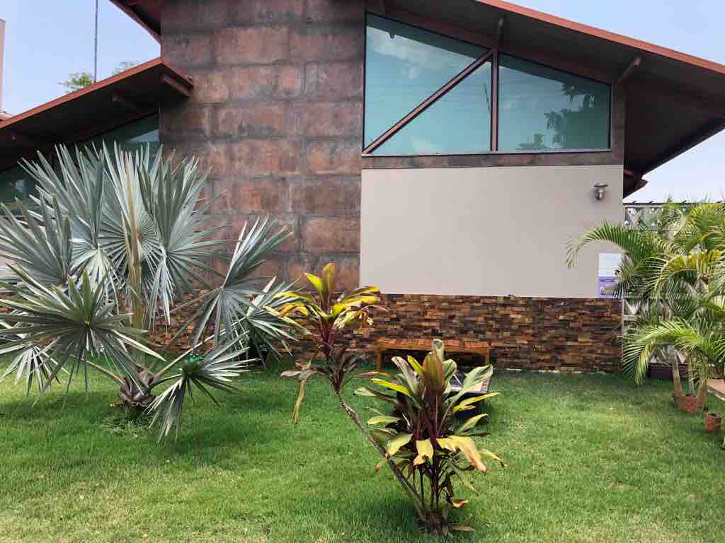 The exterior of a modern home is framed by lush greenery, featuring prominent stone walls and large glass windows. Tropical plants, including palms and colorful foliage, enhance the inviting landscape, creating a serene atmosphere in the surrounding garden.