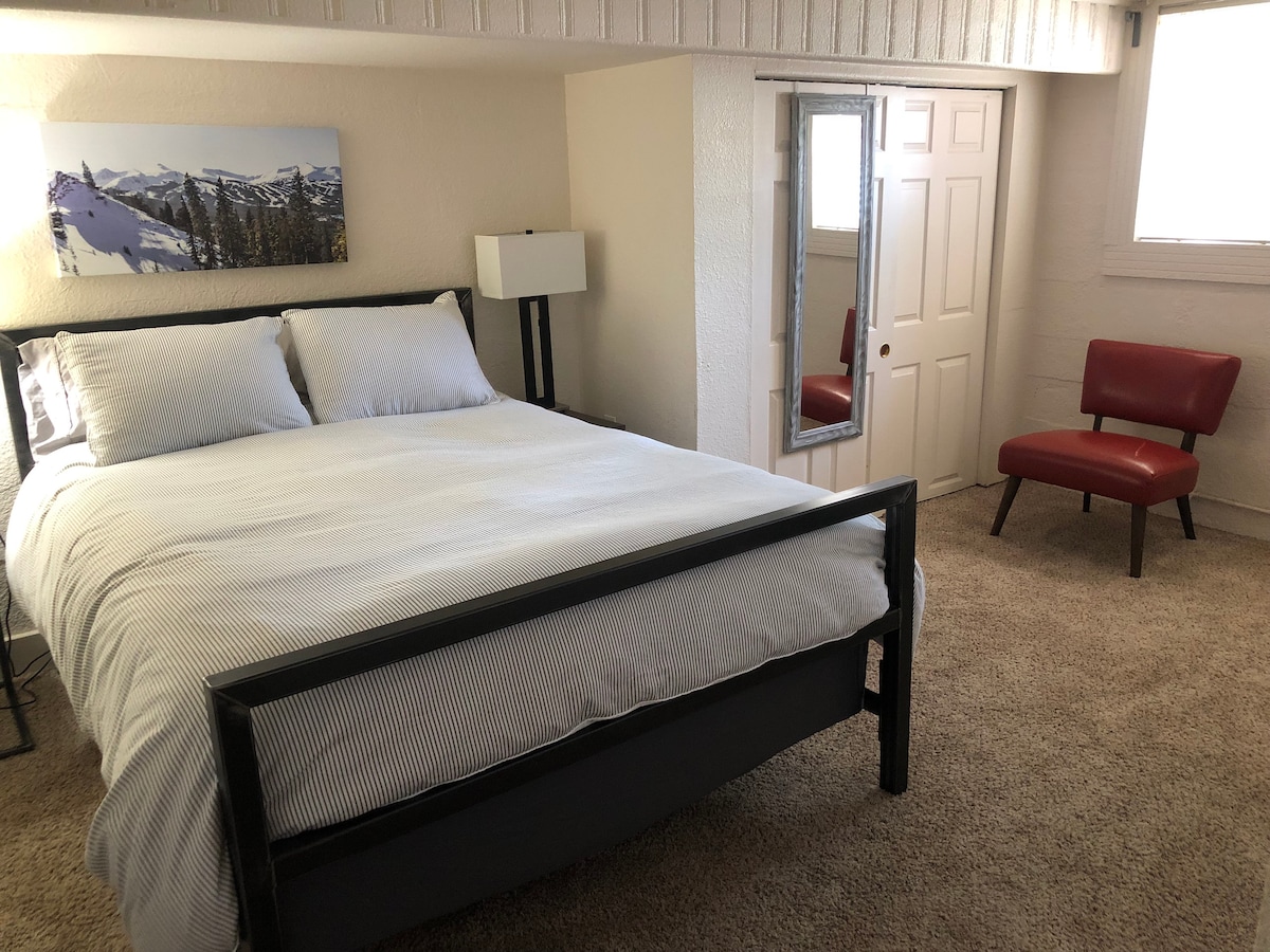 A spacious bedroom features a large bed with striped bedding, complemented by a modern lamp on the side. A red accent chair adds a pop of color, while a mirror reflects the light from a nearby window, enhancing the airy feel of the room.