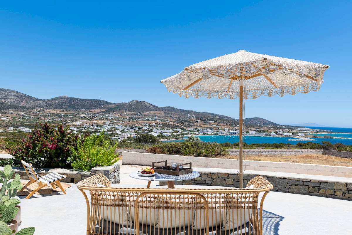 A comfortable seating area is presented outside under a decorative umbrella, overlooking Alykis' bay. The view features rolling hills and the Aegean Sea. Bright green plants and a small table with refreshments add charm to the relaxing outdoor space.