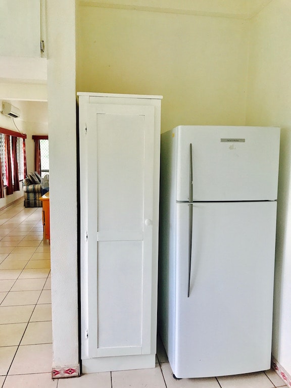 A white cupboard stands alongside a modern refrigerator, both positioned against a light-colored wall. Tile flooring extends throughout the space, with additional furnishings visible in the background, indicating an open-plan layout.