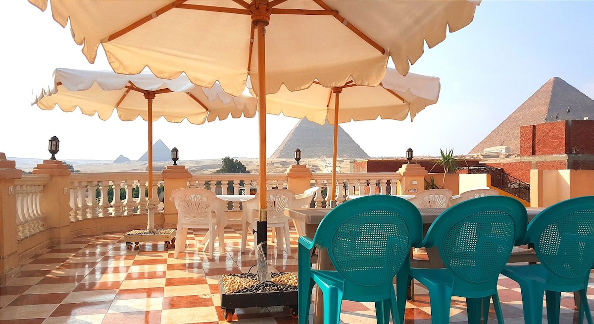 A spacious rooftop terrace features several turquoise chairs arranged around a small table. Two large umbrellas provide shade, and the backdrop showcases iconic pyramids against a clear sky. The patterned tile floor adds visual interest to the outdoor space.