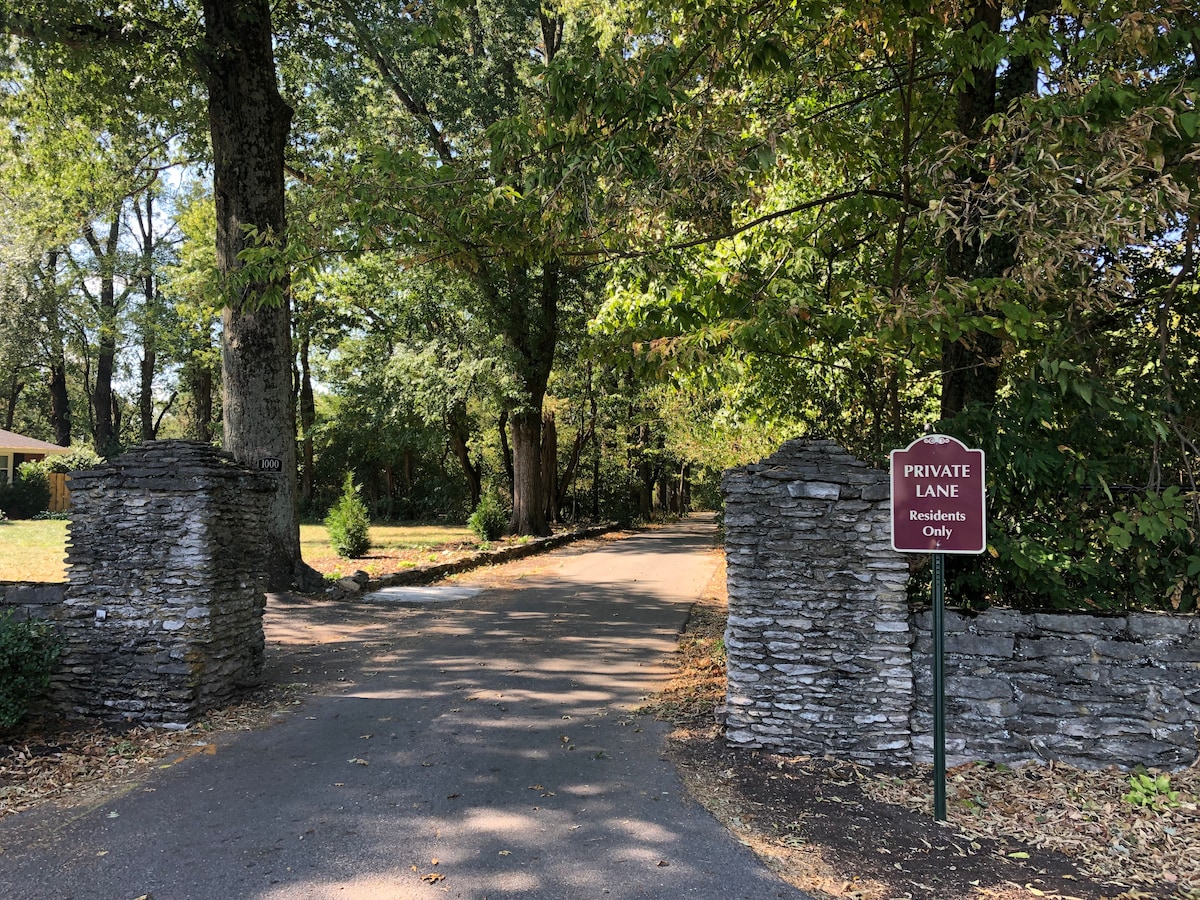 A tree-lined private lane is visible, marked by a sign indicating 'Residents Only.' The entrance is framed by sturdy stone columns and features a paved road that invites exploration into a serene, shaded area.