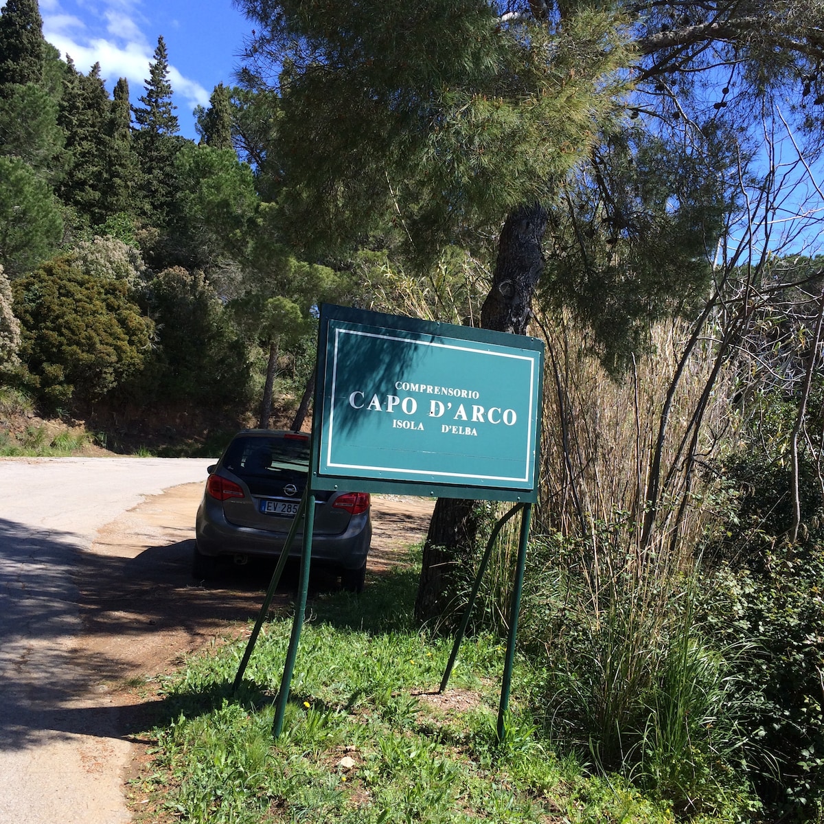 A green sign reading 'Compendio Capo d'Arco Isola d'Elba' stands alongside a dirt road, surrounded by lush trees and shrubs. A parked car can be seen nearby, indicating a welcoming entrance to the private community.