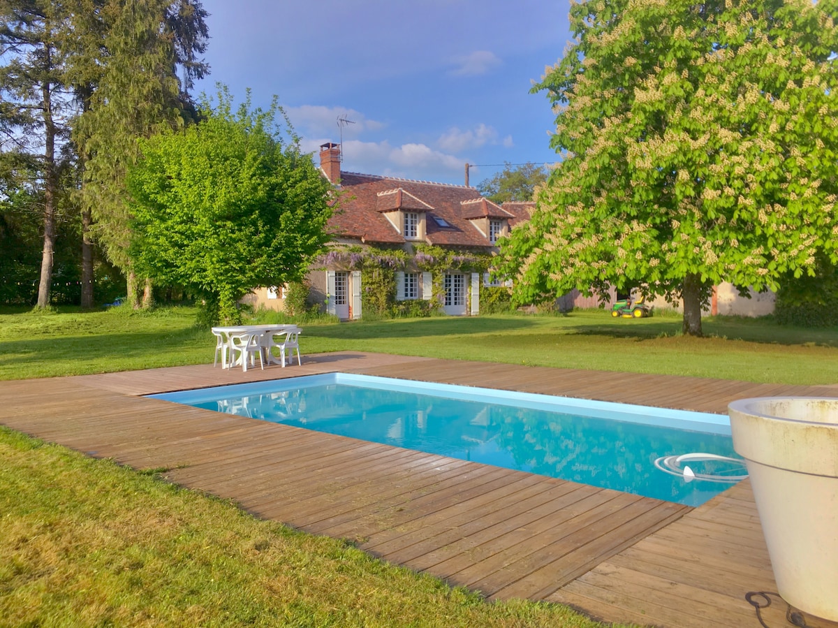 A rectangular swimming pool is framed by wooden decking and lush green grass. A dining set with white chairs and a table is positioned near the pool, providing an outdoor seating area. The house, featuring multiple windows and a rustic style, sits in the background.