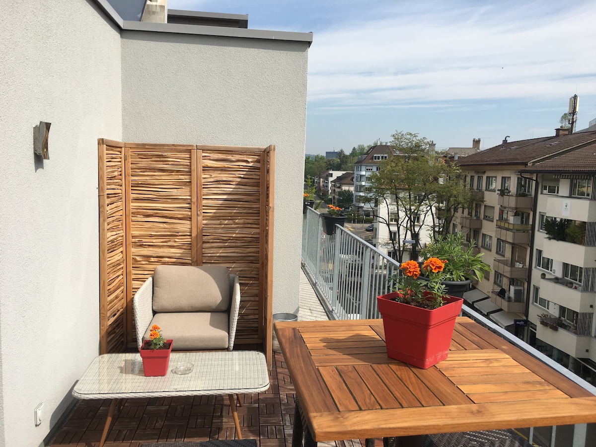 A terrace is featured, furnished with a cozy seating area and two small tables. Potted flowers add color to the space, while a wooden privacy screen provides a sense of seclusion. A view of nearby buildings and trees is visible in the background.