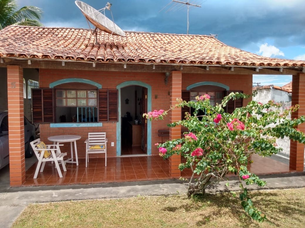 A quaint brick house is highlighted with blue window frames and wooden shutters. A small dining set with white chairs sits on the porch, surrounded by green foliage and colorful flowers. A satellite dish is mounted on the roof, with a clear sky in the background.