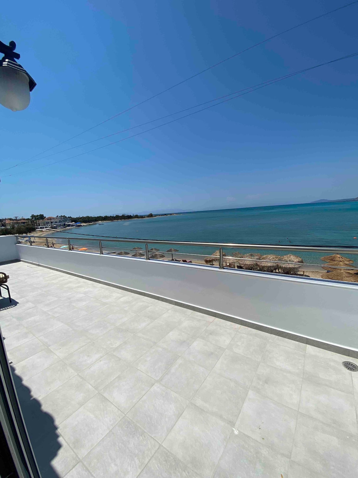 A spacious balcony offers views of the turquoise sea and sandy beach. Comfortable seating is positioned near the railing, with sunlight illuminating the tiled floor. The horizon stretches towards the blue sky, enhancing the sense of tranquility.
