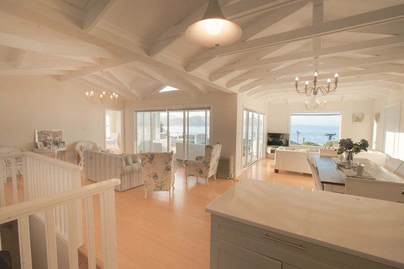 An open-plan living space features high, intricately designed ceilings and large windows that allow natural light to fill the area. Comfortable seating is arranged thoughtfully around the room, complementing the serene ocean view visible through the glass doors. A dining area is positioned to the side.