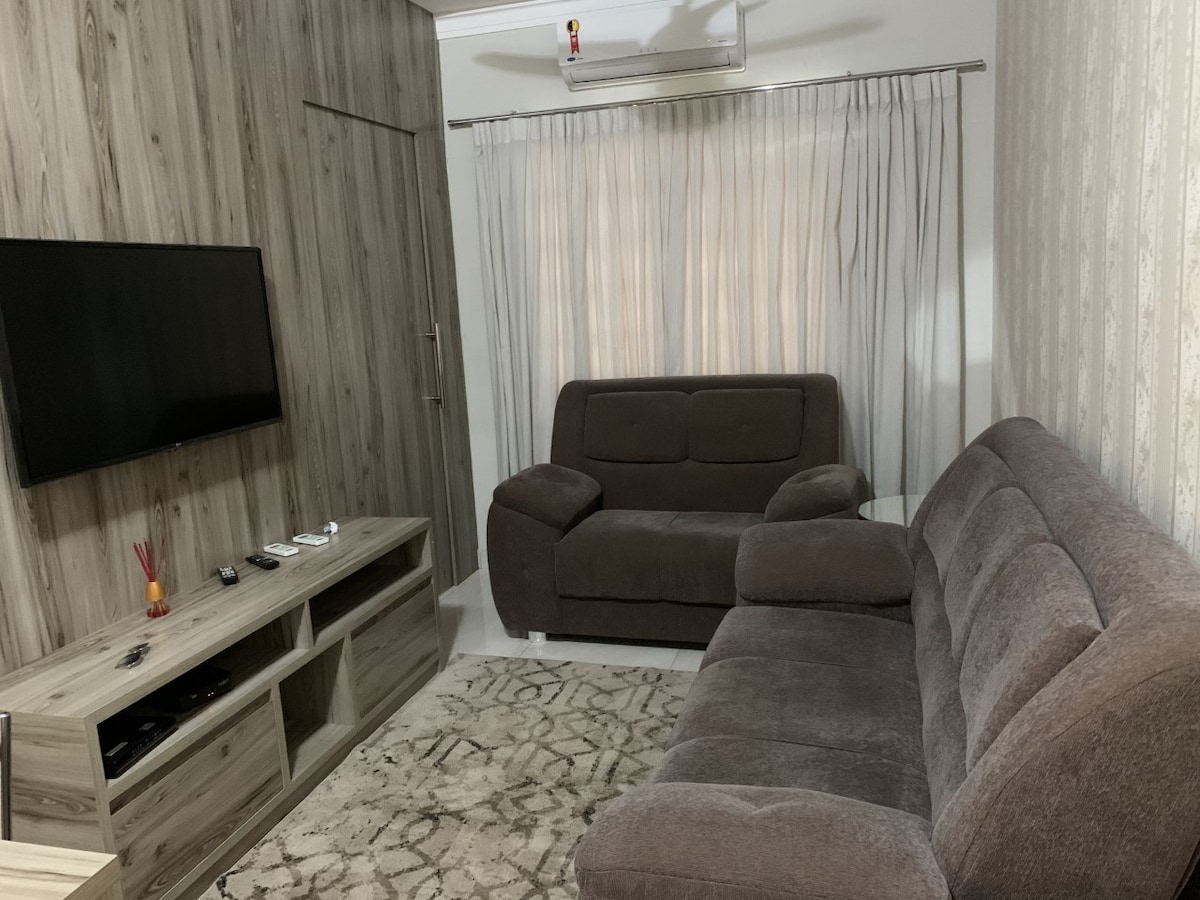 A comfortable living area is equipped with a large, soft sofa and a recliner, positioned around a light-colored entertainment unit. A wall-mounted television is visible, and soft curtains filter natural light from a nearby window, creating a calming atmosphere.