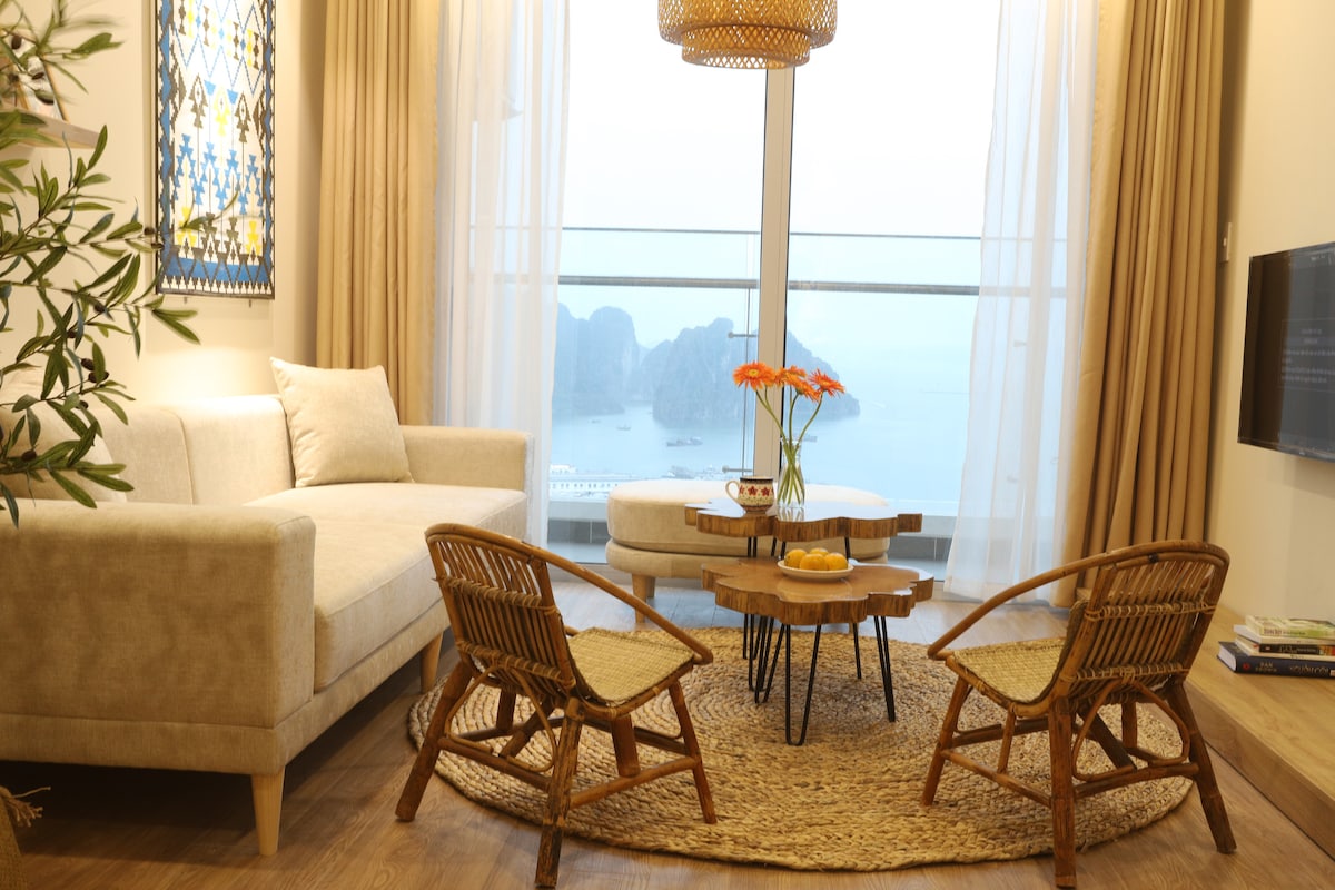 A cozy seating area features a light-colored sofa complemented by two woven chairs. A small wooden coffee table is set on a round jute rug, adorned with a bowl of fruit. Natural light floods the space, enhancing the view of the bay through large glass doors.