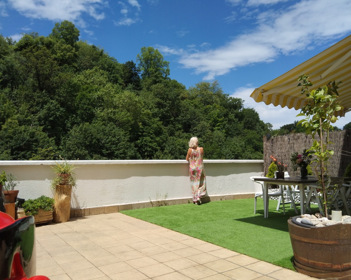A spacious terrace is shown, featuring artificial grass and tiled flooring. Potted plants add greenery around. A dining table and chairs are positioned under a striped awning. The view extends to lush trees under a blue sky, creating a serene outdoor space.