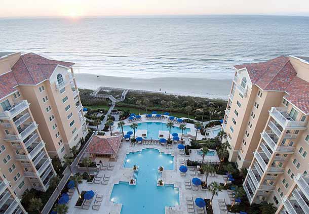An aerial view captures the oceanfront resort's expansive swimming pools, surrounded by lounge chairs and umbrellas. The sandy beach and ocean are visible in the background, with the sun setting on the horizon, casting a soft glow over the scene.