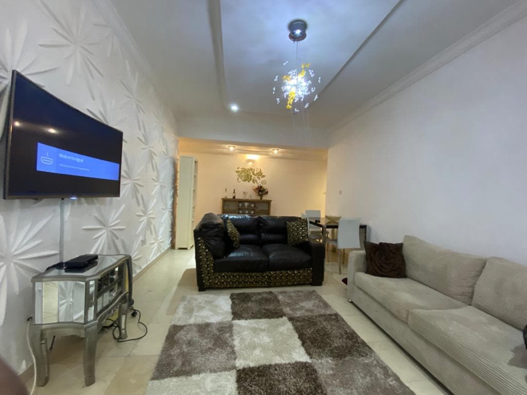 A thoughtfully arranged living area features a large TV mounted on the wall, complemented by a mix of sofas and an elegant coffee table. The textured wall adds visual interest, while a decorative light fixture illuminates the space. A soft area rug is placed on the tiled floor.