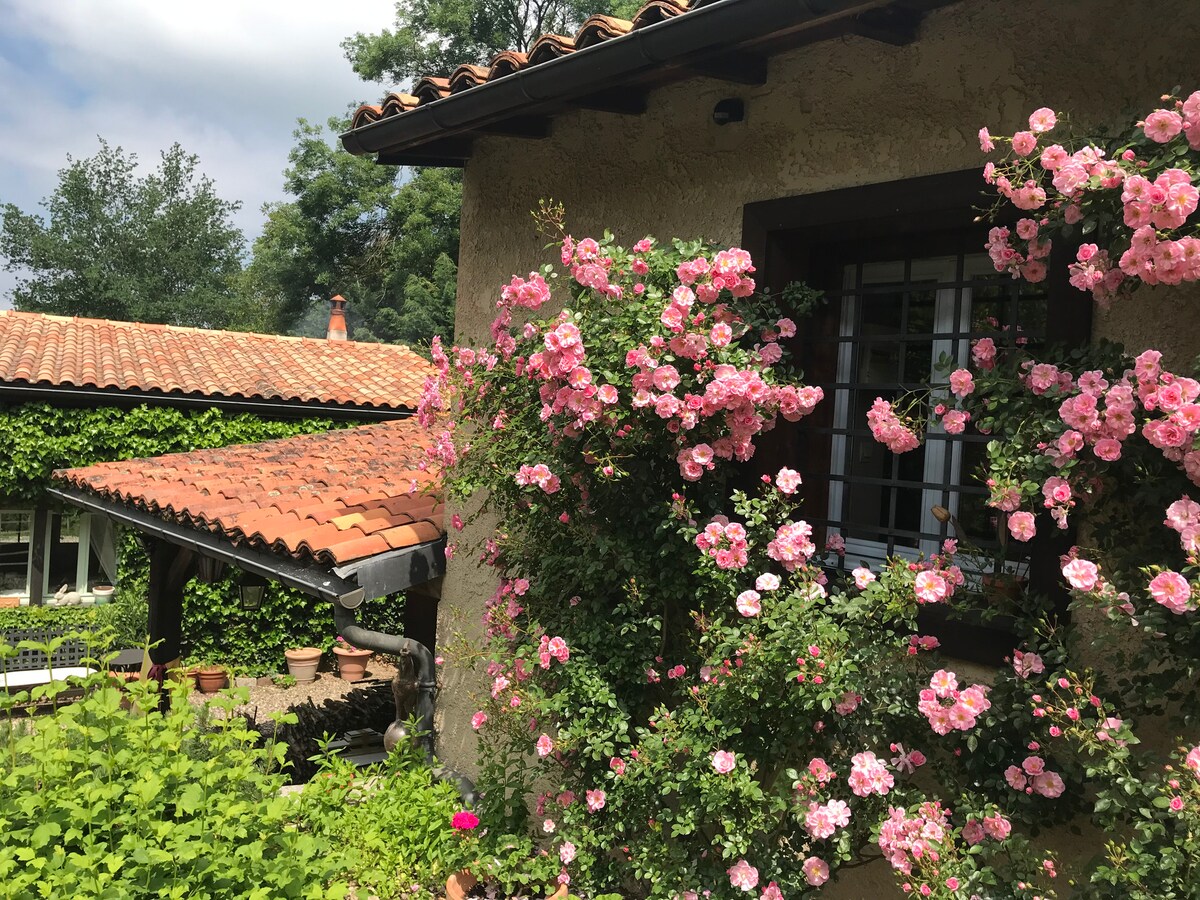 A charming exterior features a stone wall adorned with blooming pink roses, framing a window. The roof is covered with terracotta tiles, complementing the lush greenery surrounding the area. The peaceful garden space is visible in the background, offering a tranquil atmosphere.