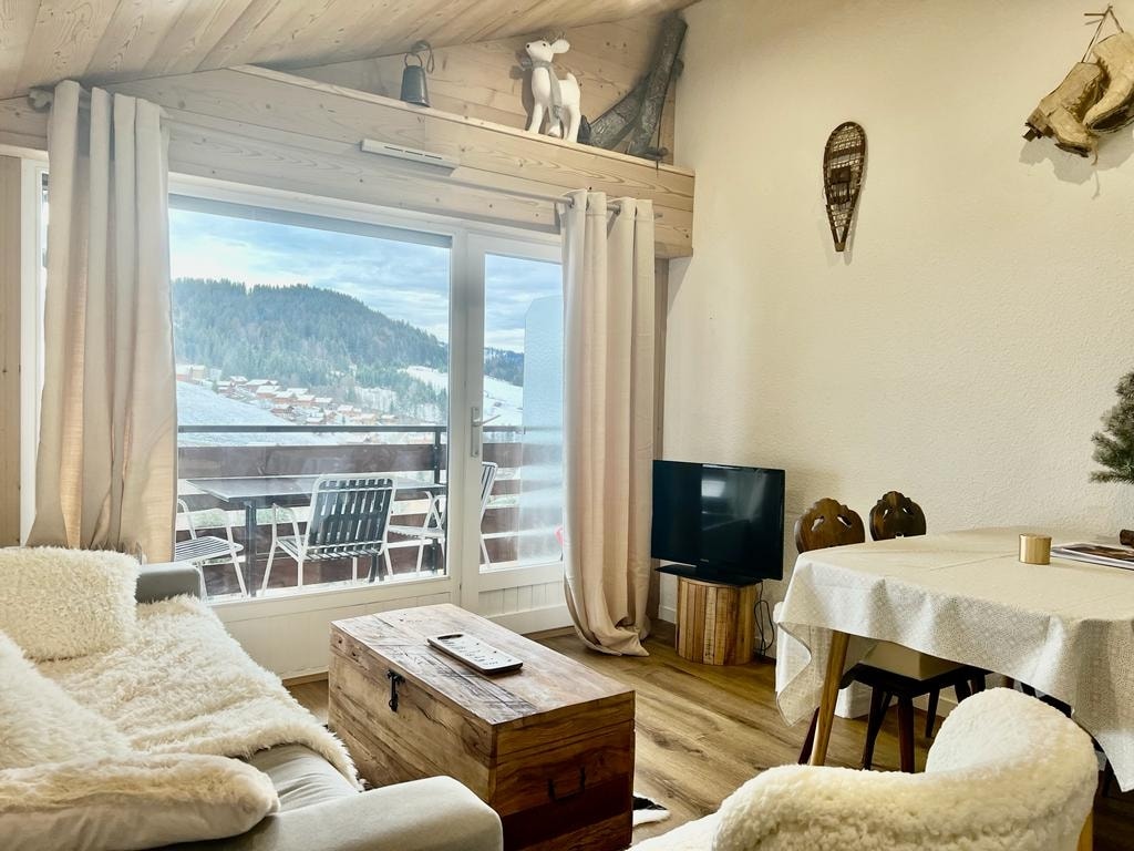 A cozy living area features a soft sofa and a wooden coffee table, complemented by a television. Large windows offer a view of the outdoor terrace, framed by soft curtains. Decorative elements include snowshoes and rustic sculptures, enhancing the mountain ambiance.