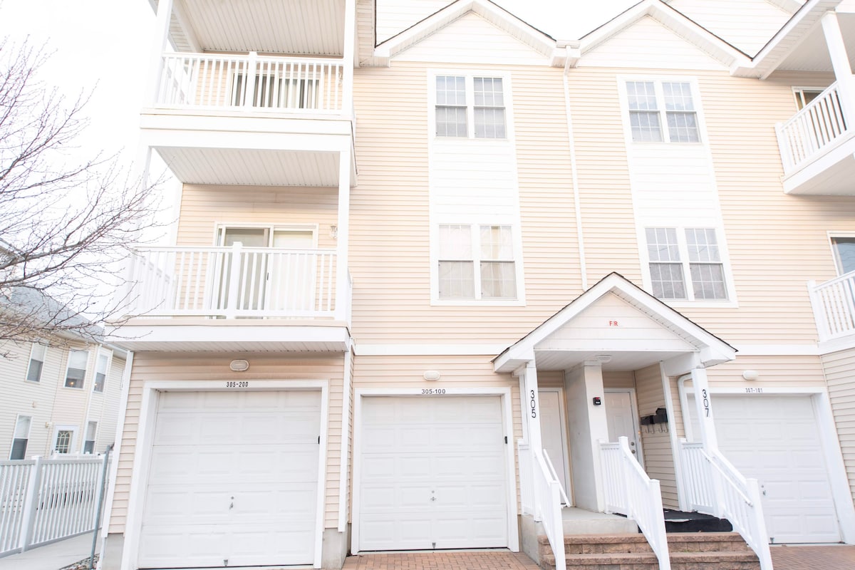 The exterior of a multi-story condo is presented, showcasing a light-colored façade with a balance of windows and white railings. A welcoming entrance features a small set of stairs leading to the door, while garages are visible on the ground level.