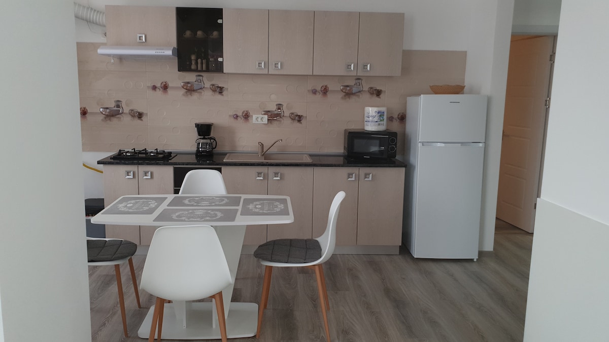 A modern kitchen area features light-colored cabinetry and a black countertop. A dining table with white chairs is positioned in front of a wall with decorative accents. Appliances including a refrigerator, microwave, and coffee maker are visible, suggesting a functional and welcoming space.