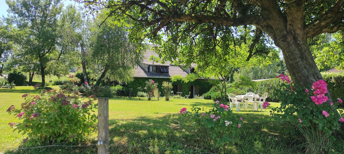 A spacious garden is surrounded by lush greenery and blossoming flowers. The house, partially covered in greenery, sits in the background. Outdoor seating is provided by white chairs, inviting relaxation amidst the natural beauty of the countryside.