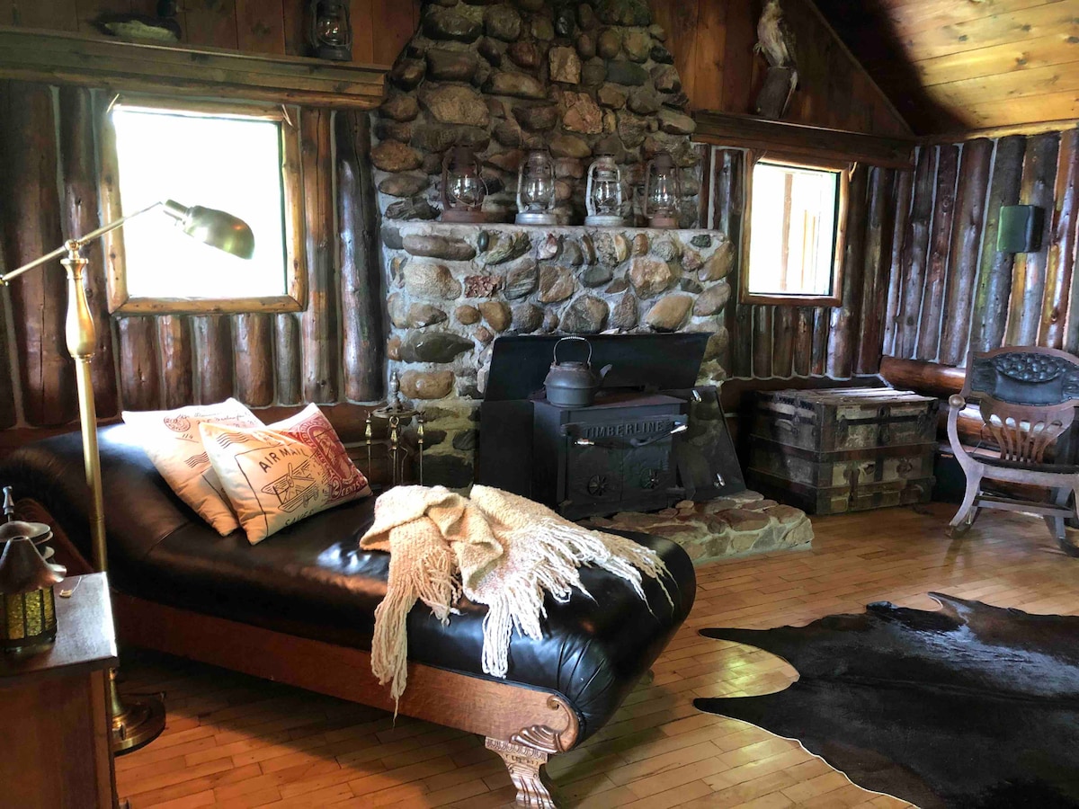 A rustic living area showcases a dark leather chaise lounge paired with a soft throw blanket. A stone fireplace serves as a central feature, flanked by wooden walls and several windows allowing natural light. A vintage chest and a wooden rocking chair add to the cozy ambiance.