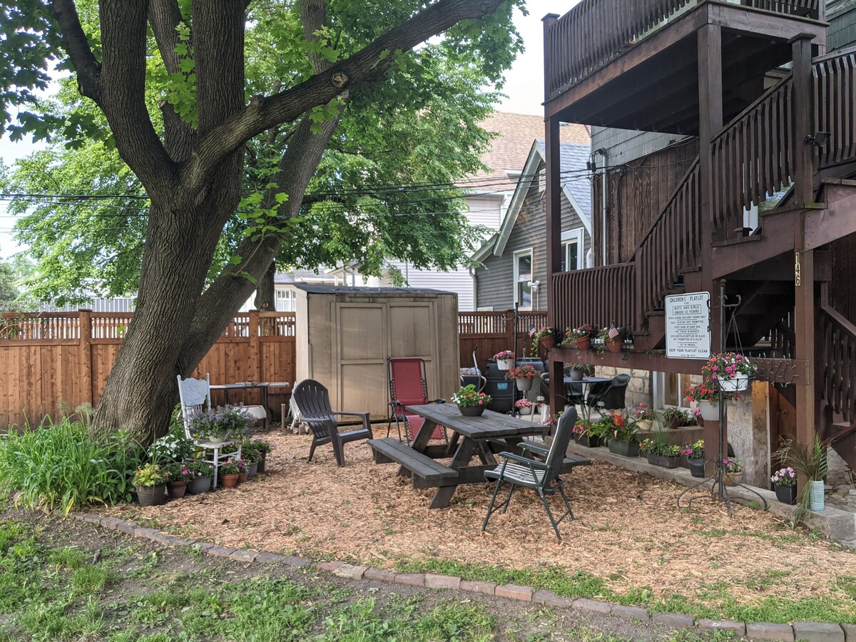 A spacious backyard area features a wooden picnic table surrounded by several chairs. Potted plants and flowers are arranged throughout, adding color to the environment. A large tree provides shade, and a storage shed resides behind the seating area, enhancing outdoor enjoyment.