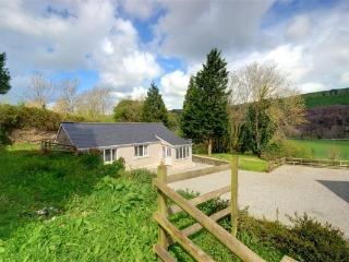 A single-story cottage is set against a backdrop of lush greenery, surrounded by trees and rolling hills. A gravel driveway is seen leading to the entrance, while a wooden fence frames the tranquil outdoor space.