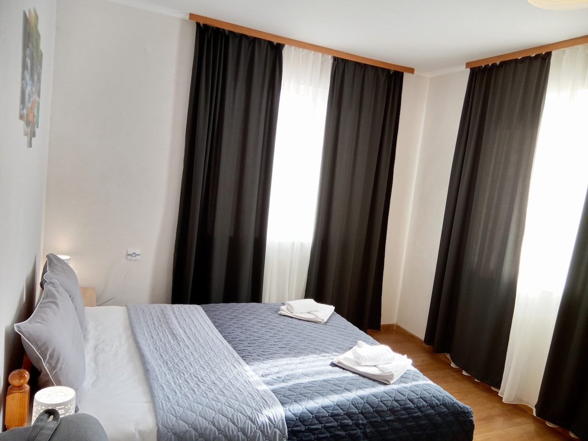 The sleeping area features a spacious bed with gray bedding and neatly folded towels. Natural light enters through large windows, which are framed by contrasting dark curtains. The walls are painted in neutral tones, contributing to a calm and relaxing atmosphere.