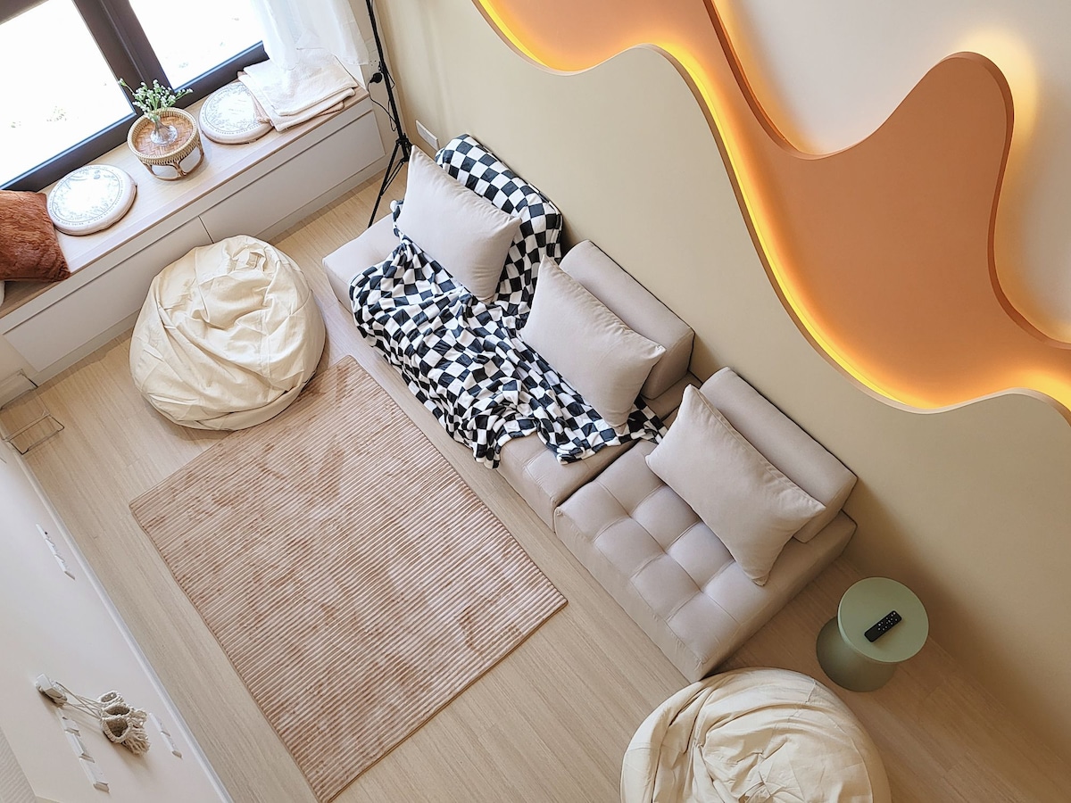 The spacious loft area features a modern sectional sofa adorned with a checkered blanket, complemented by several light-colored bean bags. A neutral-toned area rug is positioned on the wooden floor, while natural light enters through large windows, enhancing the inviting ambiance.