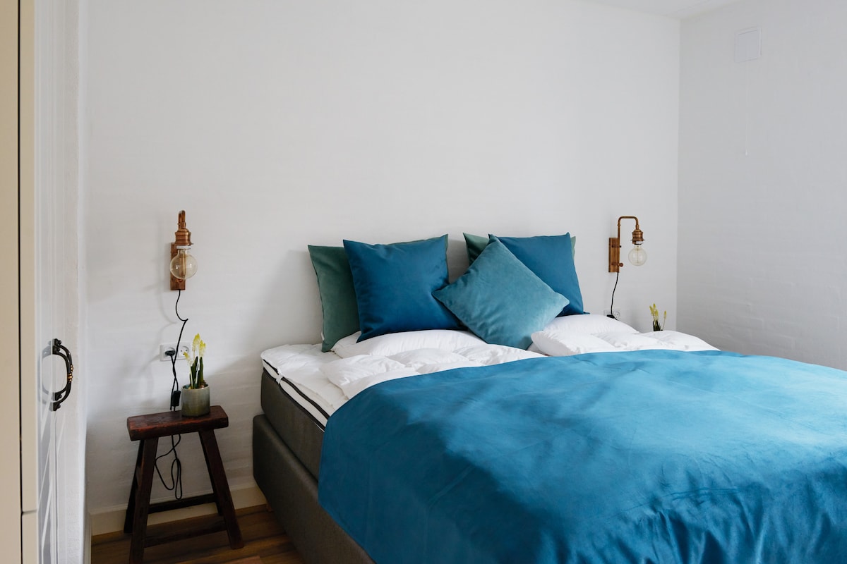 A serene bedroom features a spacious bed adorned with a deep blue coverlet and decorative pillows. Warm lighting is provided by wall-mounted lamps on either side. A small table with a plant is positioned beside the bed, creating a calm and comfortable environment.
