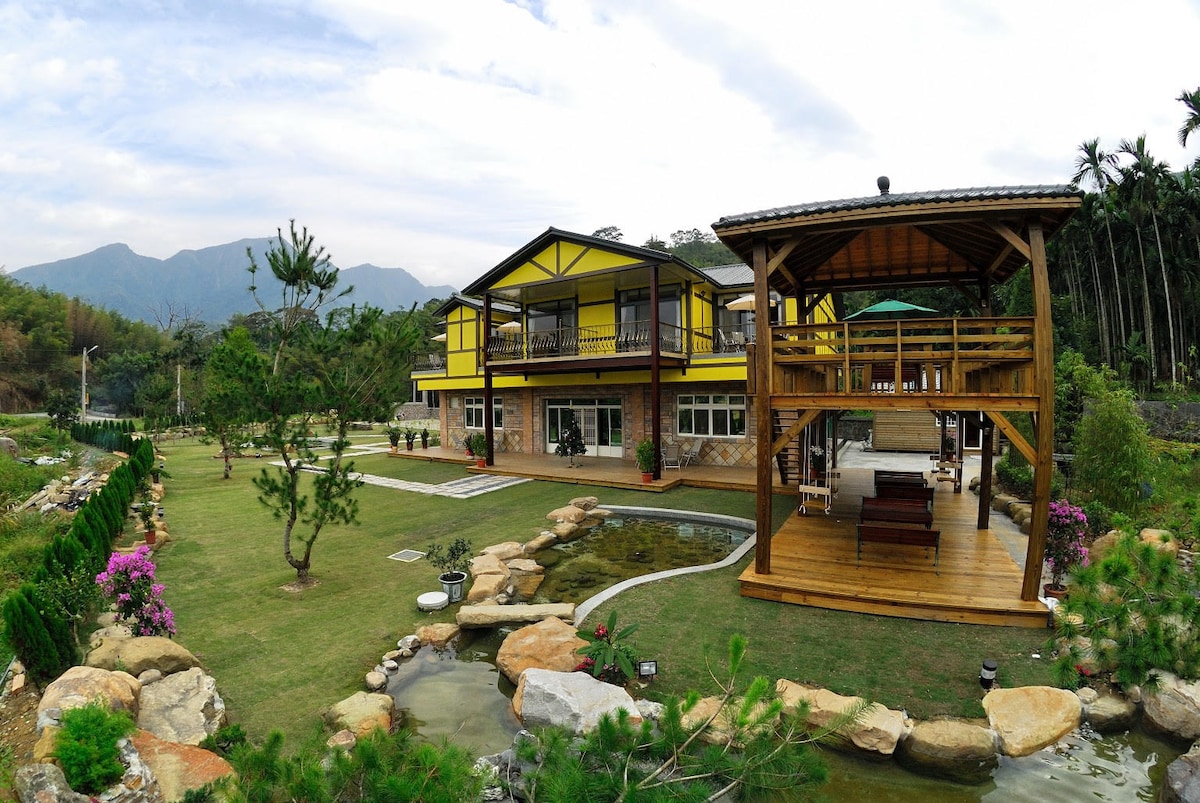 A spacious two-story building features yellow accents and large windows, surrounded by a landscaped garden with grass and ornamental rocks. A wooden gazebo is visible, providing seating areas overlooking a tranquil water feature. Lush greenery and mountains form a scenic backdrop.
