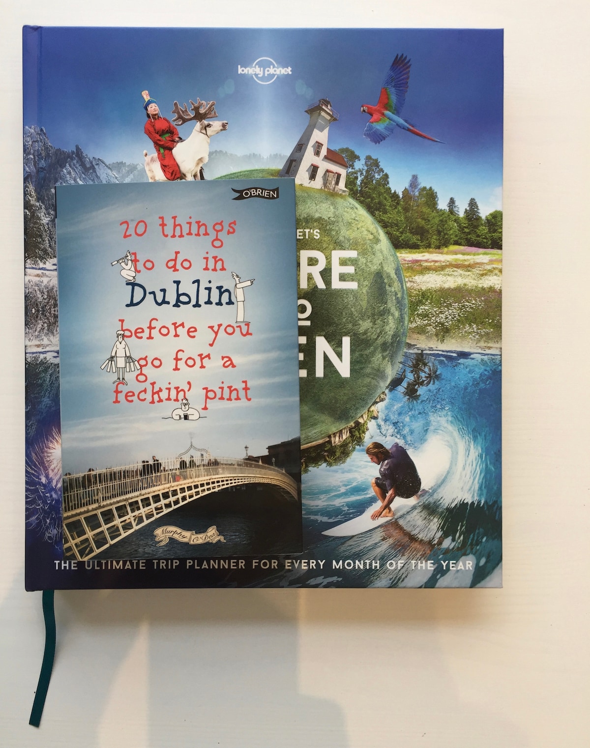 A stack of travel books and guides is displayed, featuring a prominently placed guide titled '20 things to do in Dublin before you go for a feckin' pint.' The background includes a vibrant cover of 'Lonely Planet's Nature the Green,' showcasing various landscapes and animals.