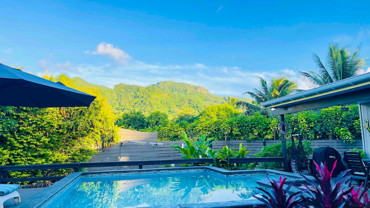 A clear view showcases the private plunge pool surrounded by lush tropical foliage. In the background, verdant mountains rise under a bright blue sky, creating a serene setting. The pool area features sun loungers and an umbrella, enhancing the space for relaxation.