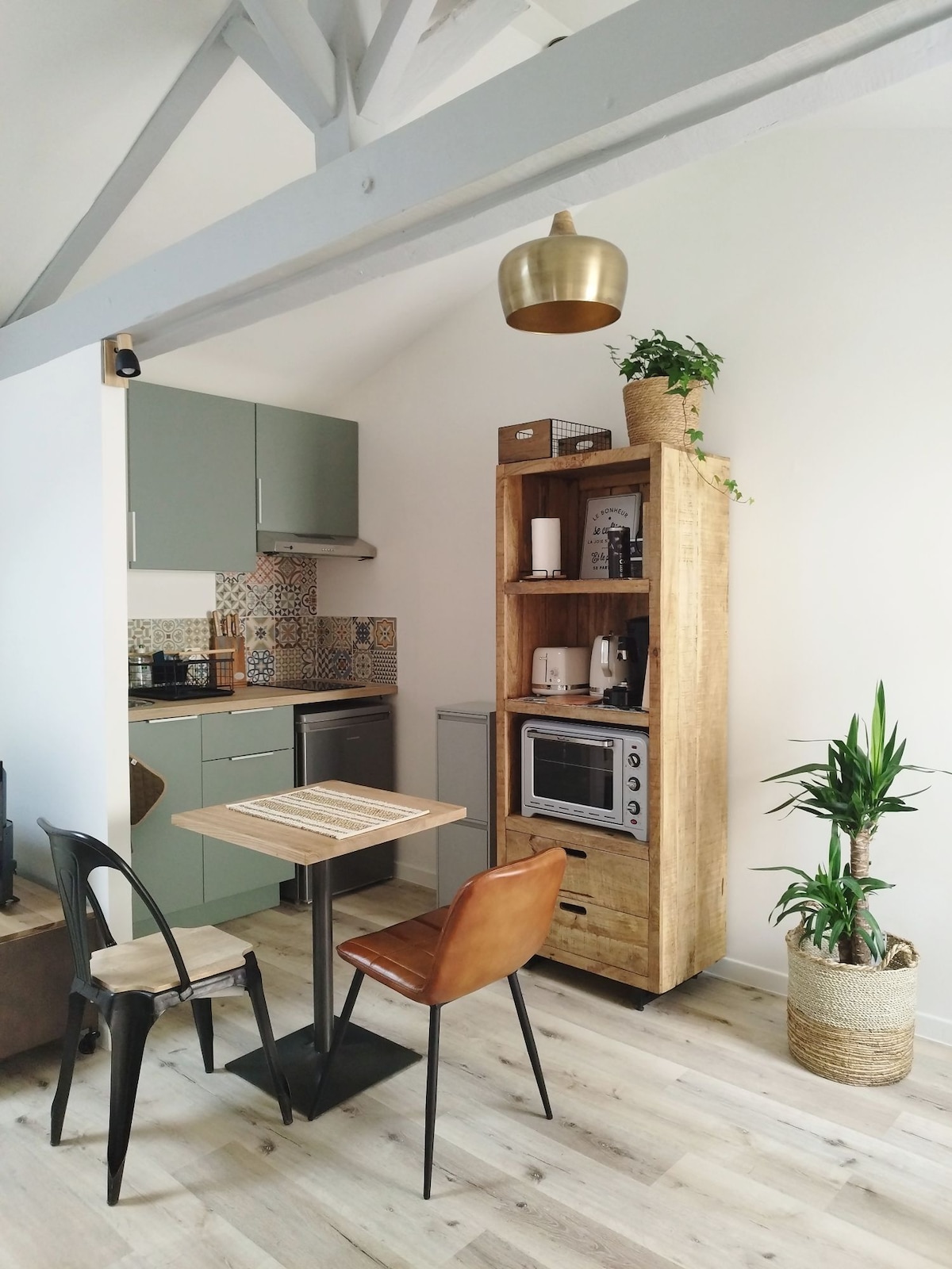 A compact kitchen area features sleek green cabinets and a wooden shelving unit, showcasing kitchen appliances and decorative plants. A small dining table with two chairs provides a cozy dining space. Light wood flooring and a high ceiling with wooden beams enhance the room's spacious feel.