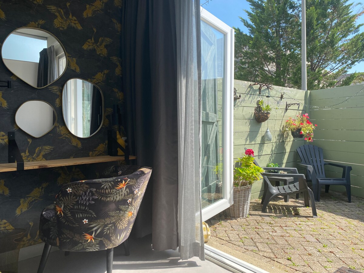 A cozy corner features a desk with decorative round mirrors above it. Large glass doors open to a private patio, where two comfortable chairs are positioned. Lush greenery and blooming flowers add a colorful touch to the outdoor space.